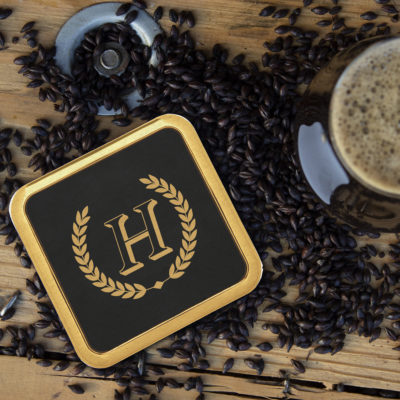SQUARE LEATHERETTE COASTER WITH METAL EDGE ON TOP OF COFFEE BEANS NEXT TO CUP OF COFFEE